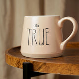 Pink earthenware mug with tapered shape, "BE TRUE" in black lettering, curved handle and bare clay base band, shown on a wooden side table.