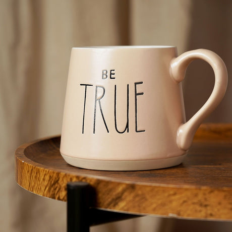 Pink earthenware mug with tapered shape, "BE TRUE" in black lettering, curved handle and bare clay base band, shown on a wooden side table.
