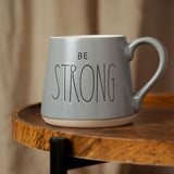 Be Strong Slogan Tapered Earthenware Mug in grey with white interior, curved handle, and engraved lettering, shown on a wooden table with a blurred room background.