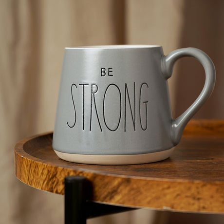 Be Strong Slogan Tapered Earthenware Mug in grey with white interior, curved handle, and engraved lettering, shown on a wooden table with a blurred room background.