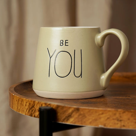 Sage green earthenware tapered mug with "BE YOU" in black lettering and a curved handle, shown on a wooden table.