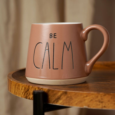 Tapered earthenware mug in matt dark taupe with white interior and "BE CALM" text, pale foot band, handle, shown on a wooden table.