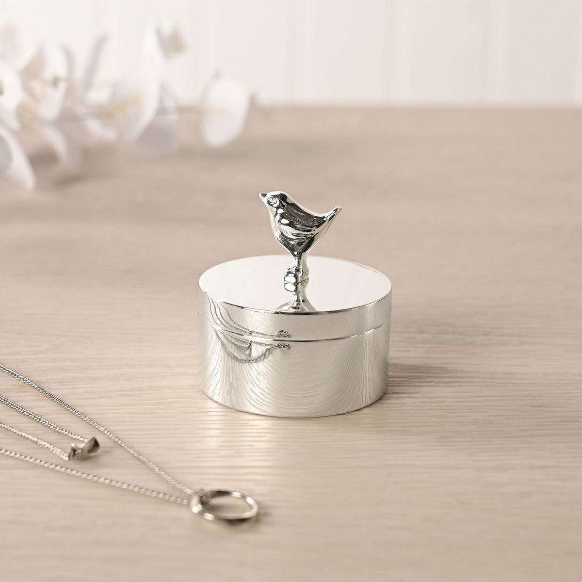 Silver-plated round trinket box with a robin figure as the lid handle, displayed on a wooden surface near a necklace and blurred white flowers.