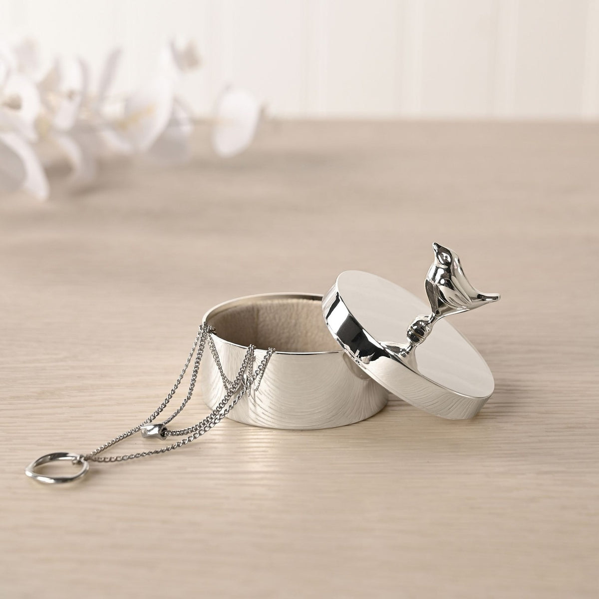Silverplated Robin Round Trinket Box with lining, lid open and robin finial, holding a necklace and ring, on a light wooden surface with white flowers behind.