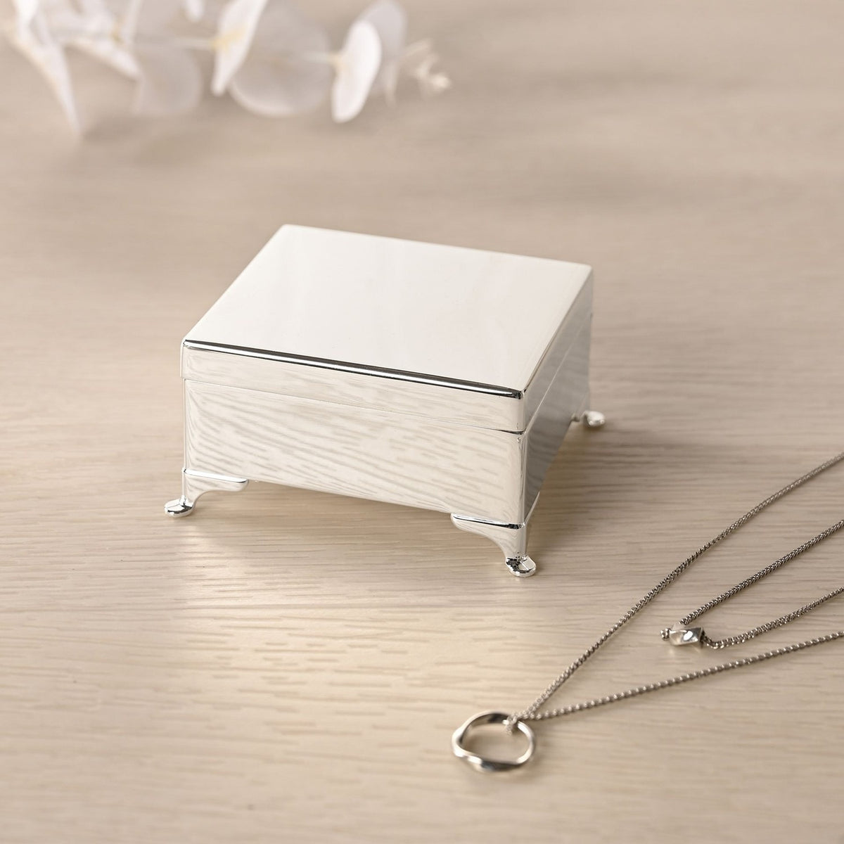 Silverplated rectangular trinket box with four feet, polished silver finish and hinged lid, shown closed on a wooden surface beside a necklace.