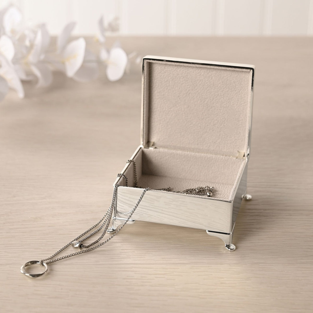 Open silverplated rectangular trinket box with four feet, polished silver finish and lined interior, holding a necklace, shown on a light surface with flowers behind.