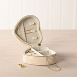 Open heart-shaped cream plastic jewellery box with pebble-grain texture, gold-tone zip, ring rolls and divided compartments holding rings and earrings, shown with a gold chain on a light wood surface.