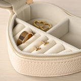 Heart-shaped cream plastic jewellery box, open to show velvet ring rolls holding a gold ring and gold stud earrings, plus a gold heart charm, with a gold-tone zip and pebble-grain exterior.