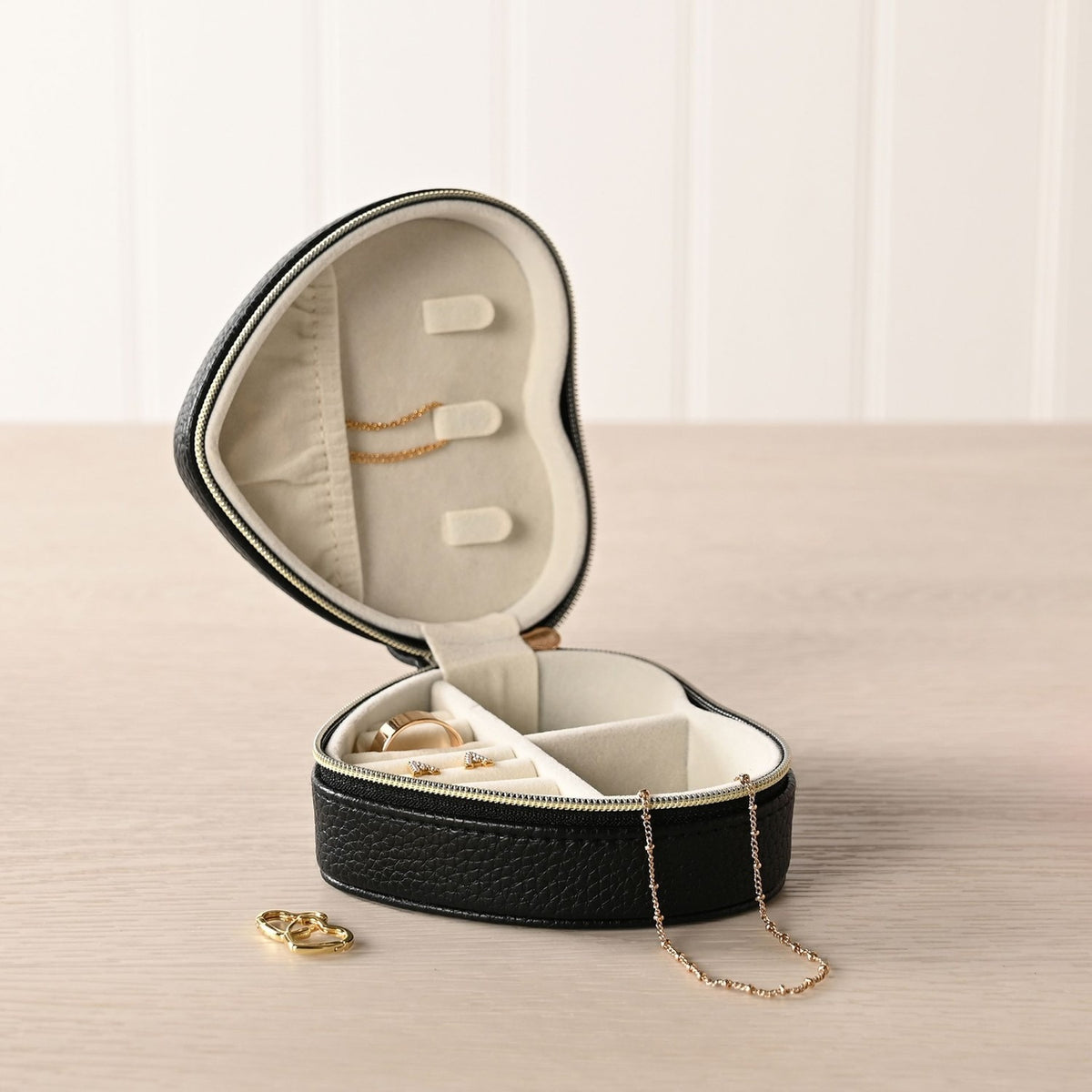Open heart-shaped jewellery box in black pebbled plastic with cream interior, ring rolls, chain holder and zip divider, shown with rings and necklaces on a wooden surface.