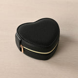 Heart-shaped jewellery box in black pebbled-grain plastic with stitched edges and a gold-tone zip with rose gold pull, shown closed on a neutral background.