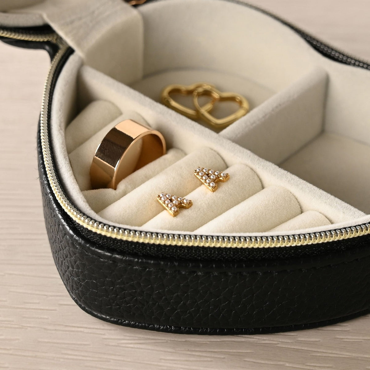 Heart-shaped jewellery box in black pebbled-grain plastic with gold-tone zip, open to show cream interior with ring rolls holding a gold ring, stud earrings, and a linked-hearts charm.