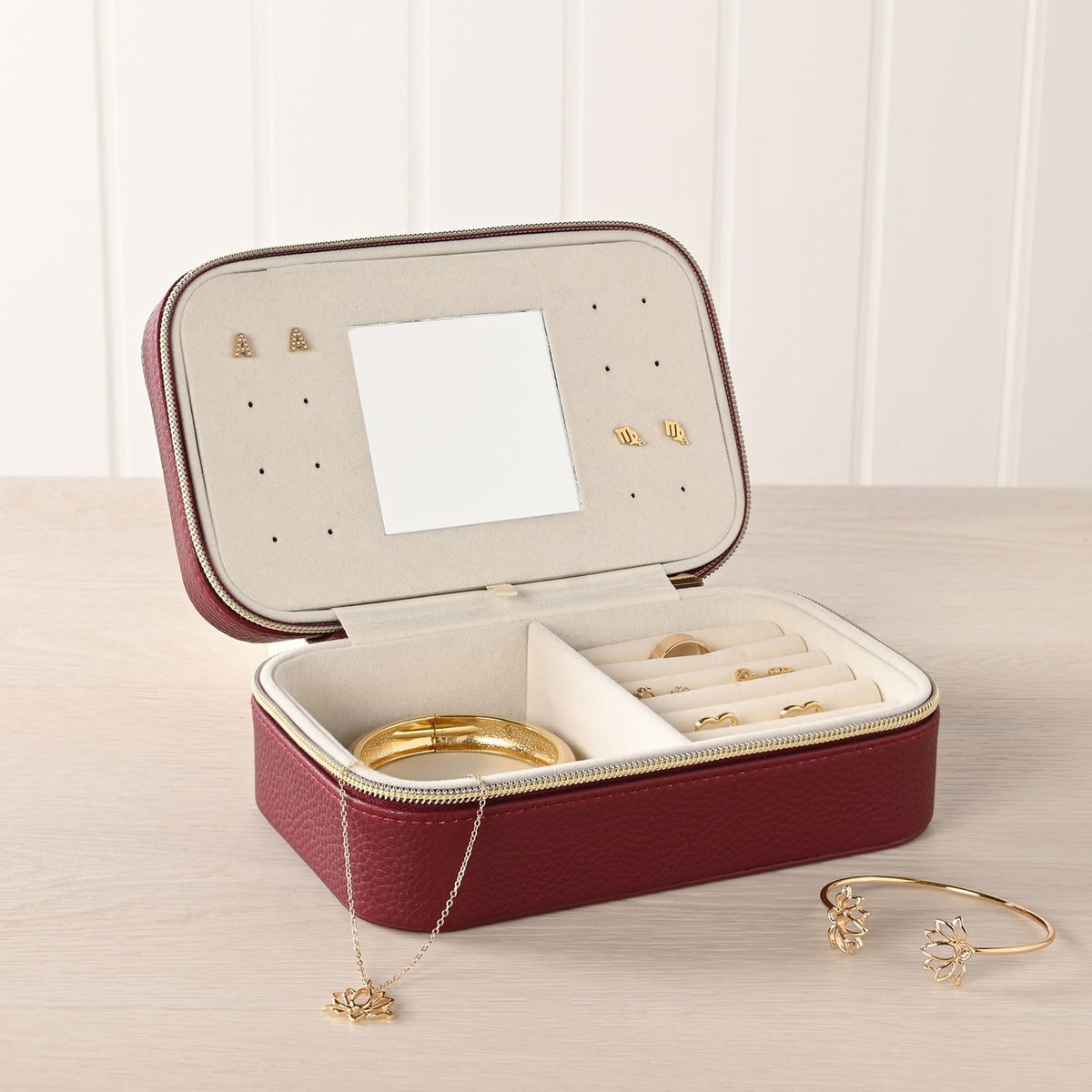 Medium rectangular jewellery box in red pebbled plastic with gold-tone zip, open to show a cream-lined interior with ring rolls, earring holes, a lid mirror, and jewellery displayed alongside.