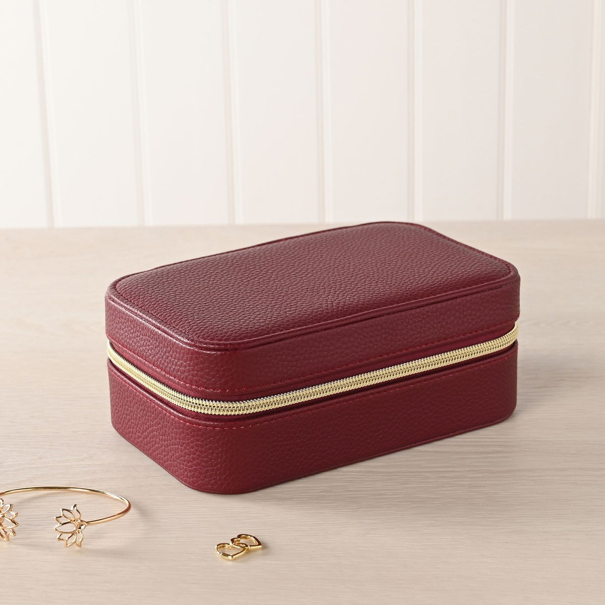 Medium rectangular jewellery box in red pebbled-texture plastic with a gold-tone zip closure, shown closed beside a gold bracelet and earring on a light wooden surface.