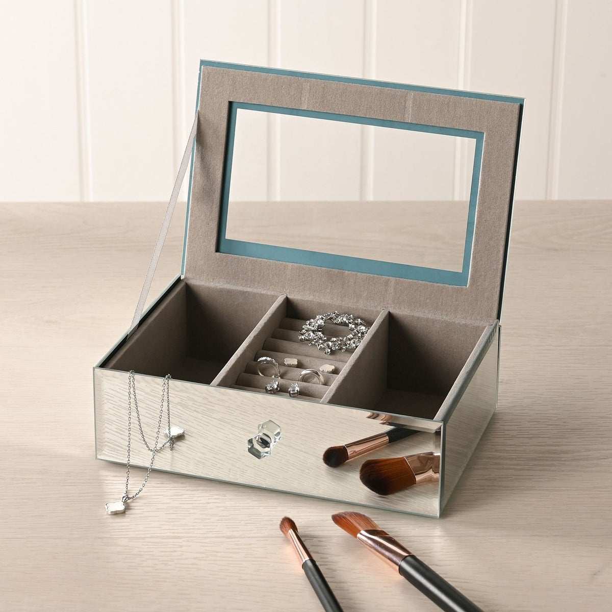 Open silver mirror glass jewellery box with taupe lined interior, central ring roll, divided compartments holding jewellery, shown on a dressing table with makeup brushes.