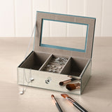 Open silver mirror glass jewellery box with taupe lined interior, central ring roll, divided compartments holding jewellery, shown on a dressing table with makeup brushes.