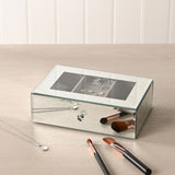 Rectangular mirror glass jewellery box in silver with glass lid revealing a taupe-lined interior and ring roll, displayed on a dressing table with brushes and a necklace.