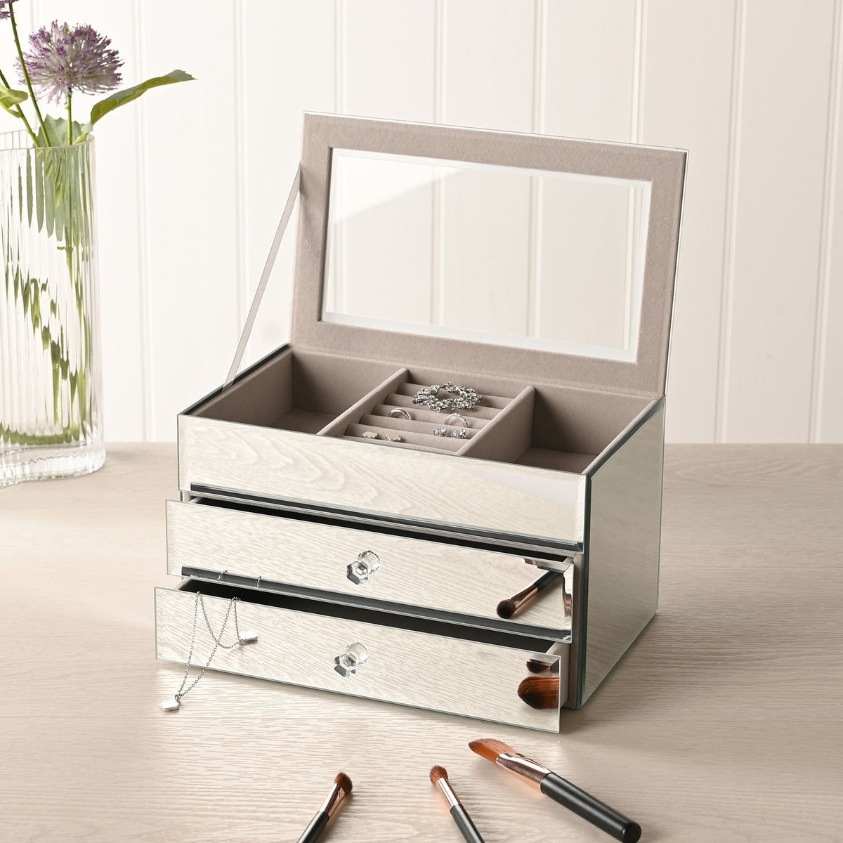 Rectangular 2 drawer mirror glass jewellery box in silver, lid open to show a lined tray with ring roll, holding rings and a necklace on a dressing table.