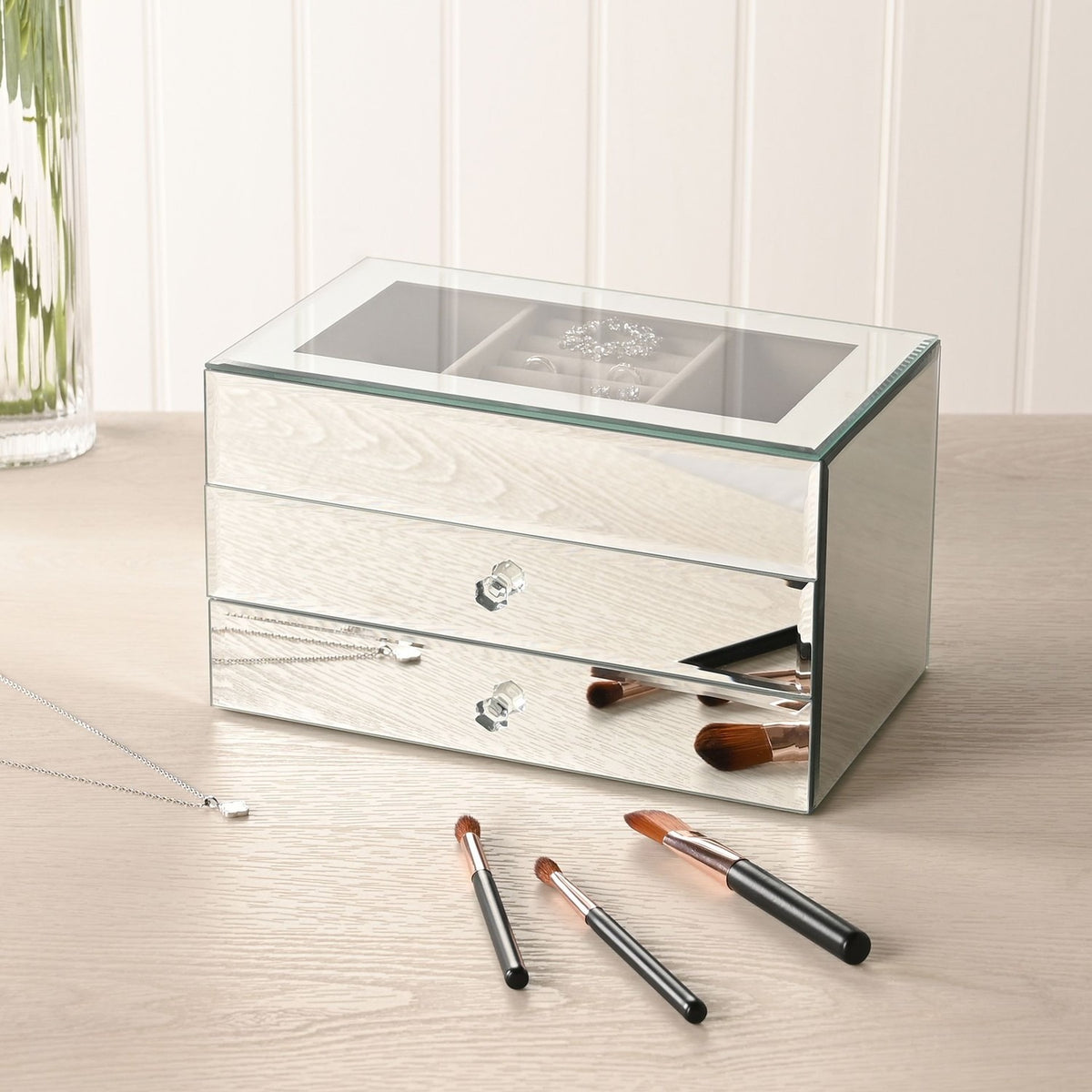 Rectangular mirror glass jewellery box in silver with two drawers open, crystal-style knobs and a lined top tray, shown on a dressing table with makeup brushes and a necklace.