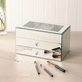 Rectangular mirror glass jewellery box in silver with two drawers open, crystal-style knobs and a lined top tray, shown on a dressing table with makeup brushes and a necklace.