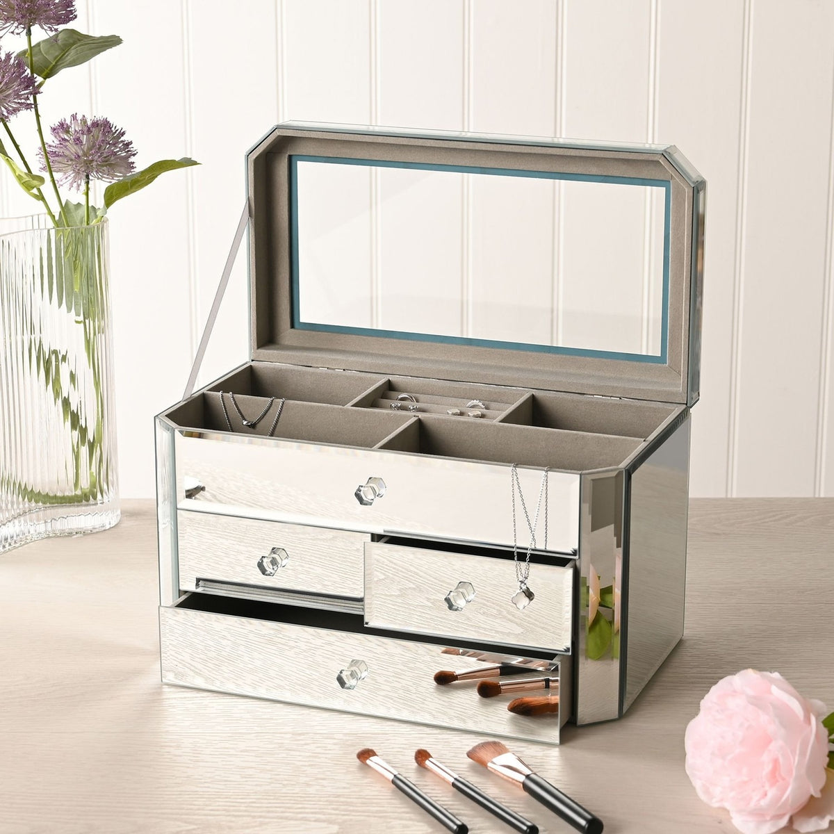 Rectangular 4 drawer mirror glass jewellery box in silver, open lid revealing a mirrored interior tray, with faceted knobs on each drawer, shown on a dressing table.