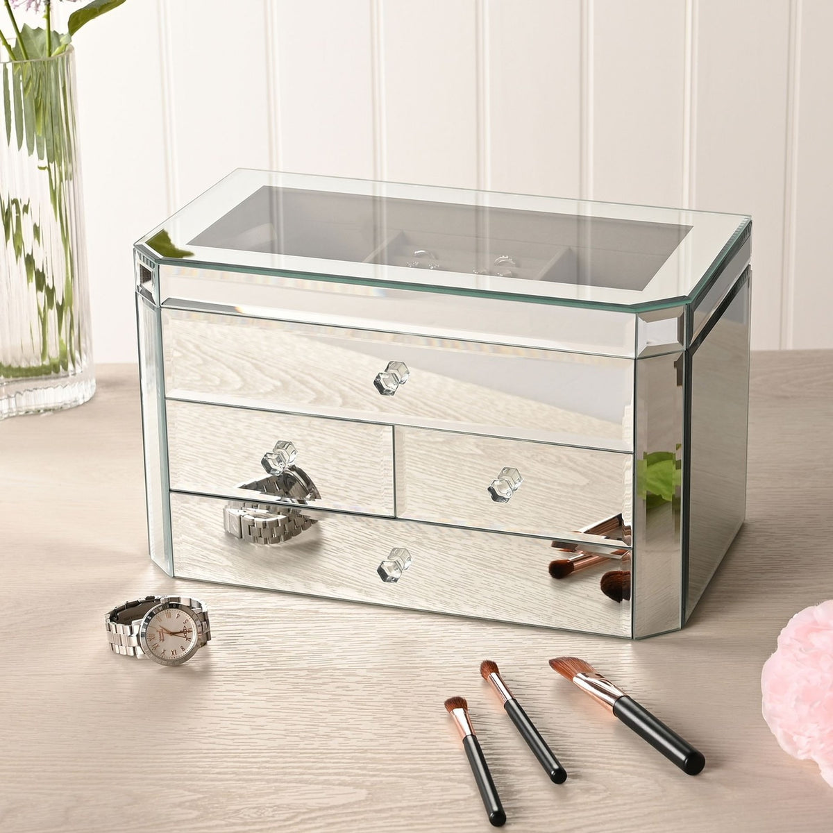 Rectangular mirror glass jewellery box with silver finish, four drawers with clear faceted knobs and a lift-up lid, shown on a dressing table with a watch and makeup brushes.