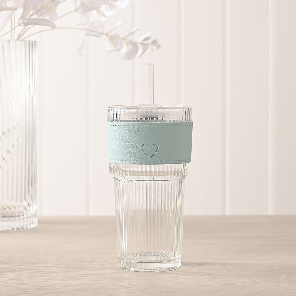 Ribbed cold drink tumbler with clear fluted glass body, blue sleeve with embossed heart, fitted lid and clear straw, shown on a wooden surface with soft decor in the background.