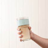 Hand holding a ribbed clear cold drink tumbler filled with a milky beverage, featuring a blue sleeve with a heart detail, clear lid and straw, against a light background.