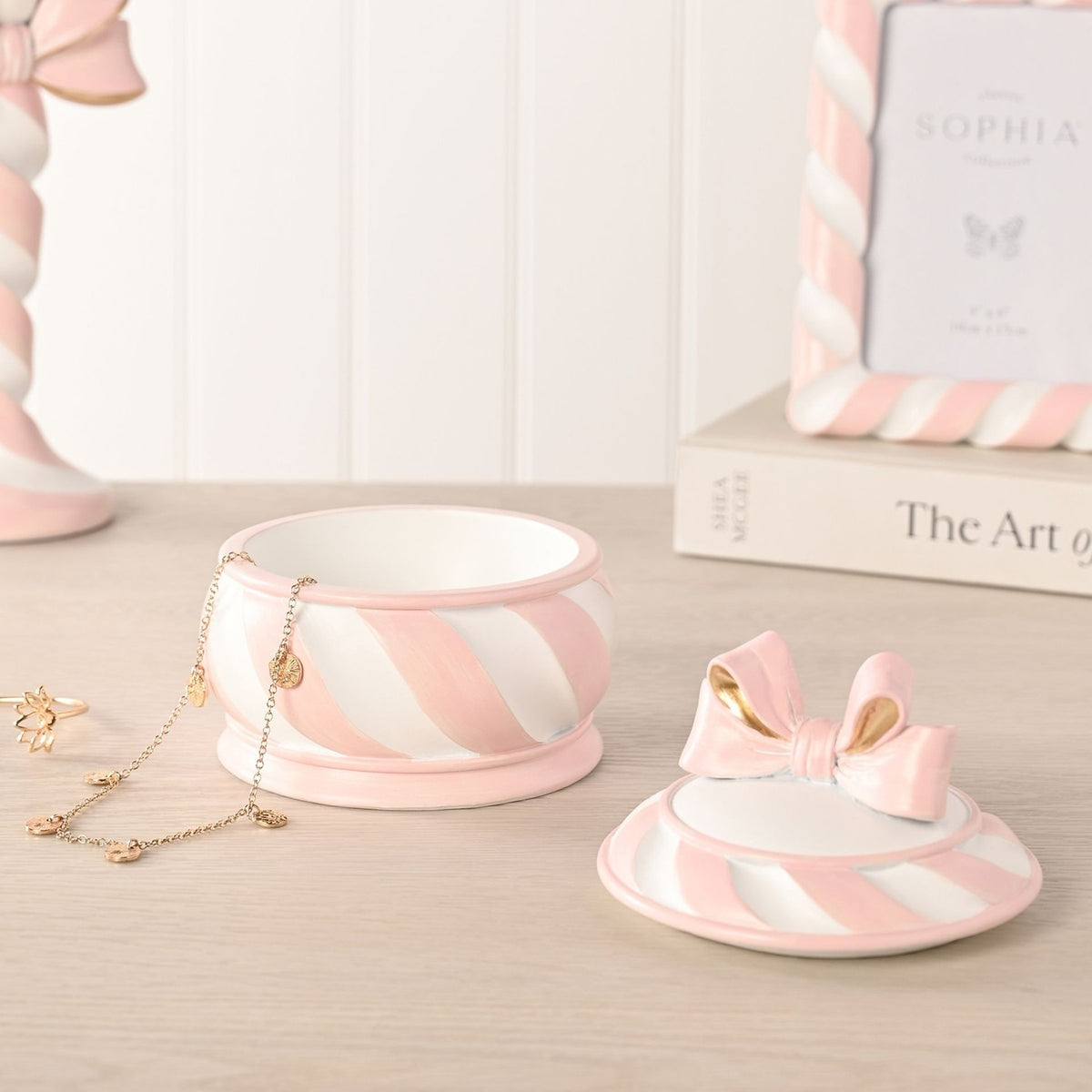 Round Candy Cane Bow Resin Trinket Box, 12cm, with pink and white striped base and gold-accented bow lid, shown open beside jewellery on a tabletop setting.