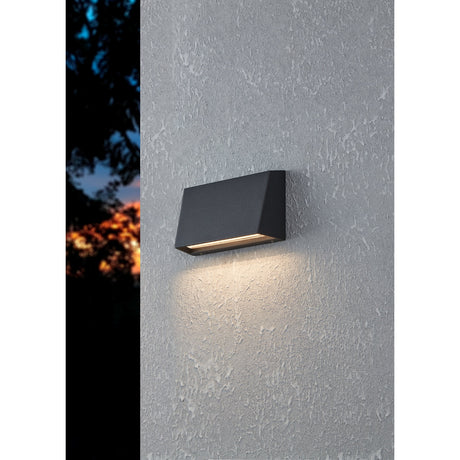 Spongano LED Outdoor Wall Light in black aluminium, wedge-shaped design mounted on a textured grey exterior wall, casting warm downward light at dusk.
