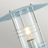 Close-up of the Stockholm Outdoor 1 Light Large Wall Lantern in a modern cylindrical design, crafted from galvanized steel with a silver finish. It showcases a clear glass casing and metal accents, with an illuminated bulb inside. Rated IP54 for durability and elegance, it stands out beautifully against a plain, light gray background.