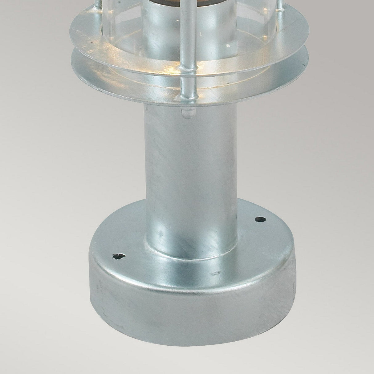 The Stockholm 1 Light Pedestal - Silver, a galvanized steel cylindrical object with a round base and layered top that resembles a miniature lighthouse or decorative post light, embodies a Scandinavian design aesthetic against a plain gray background.