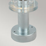 The Stockholm 1 Light Pedestal - Silver, a galvanized steel cylindrical object with a round base and layered top that resembles a miniature lighthouse or decorative post light, embodies a Scandinavian design aesthetic against a plain gray background.