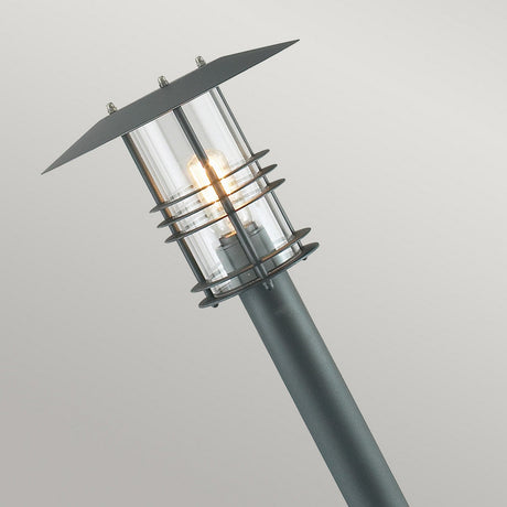 Introducing the Stockholm 1 Light Pillar - Black, a contemporary outdoor lamp featuring a galvanized steel frame and a clear cylindrical glass enclosure that highlights the visible glowing bulb within. This lamp exemplifies Scandinavian design with its sleek, angled structure set against a minimalist background.