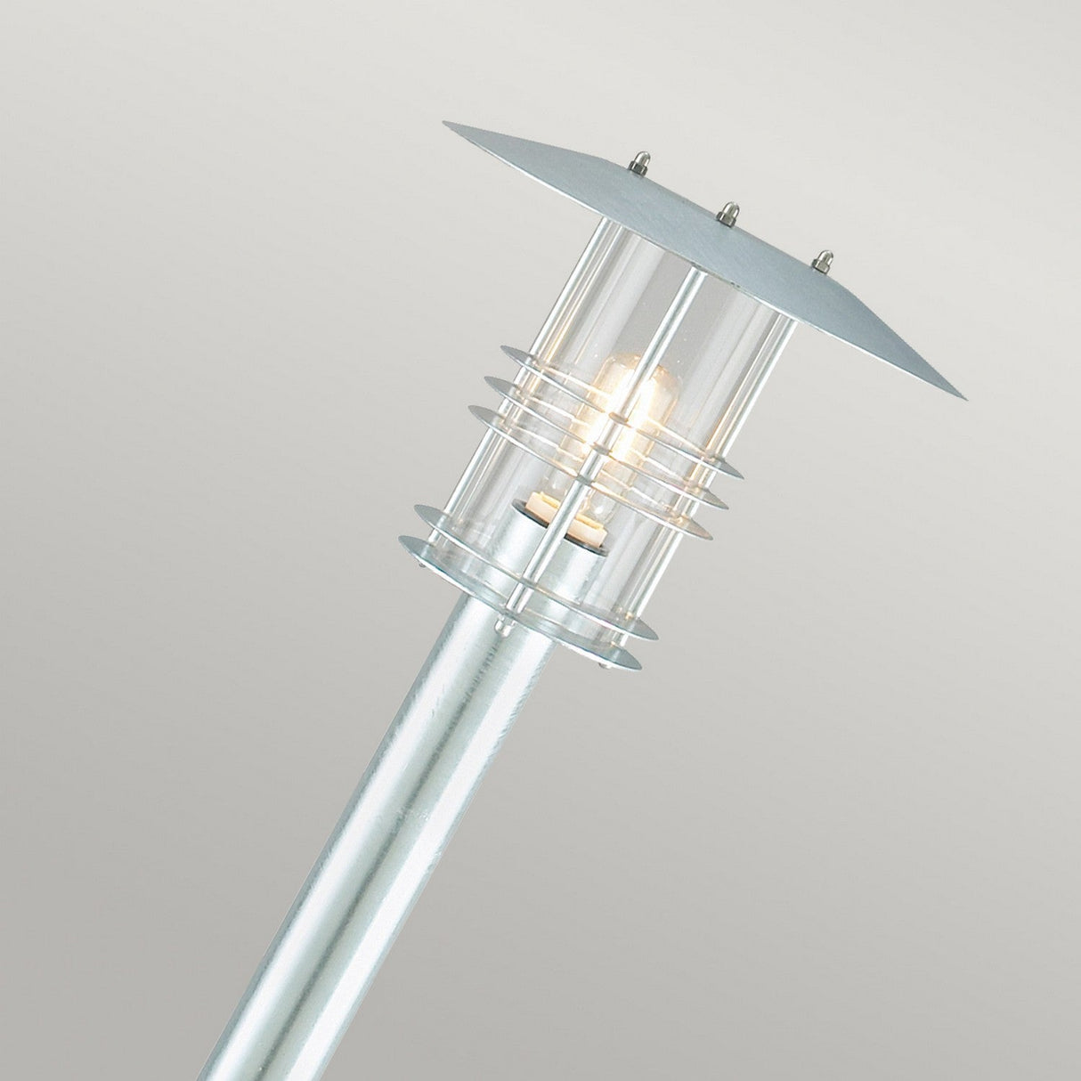 The Stockholm 1 Light Pillar - Silver features a sleek, cylindrical post made of galvanized steel with a transparent glass enclosure encasing the bulb, all topped with a flat metal cover. When illuminated, it emits a warm glow. Its minimalist design reflects Scandinavian aesthetics against a simple soft gray backdrop.