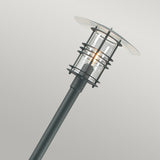 The Stockholm 1 Light Grande Lamp Post - Black features a sleek, modern design with a black pole and Scandinavian-inspired cylindrical glass enclosure that houses a lit bulb. Its flat, disc-shaped top elegantly stands out against a simple, light gray background, making it the epitome of chic outdoor lighting.