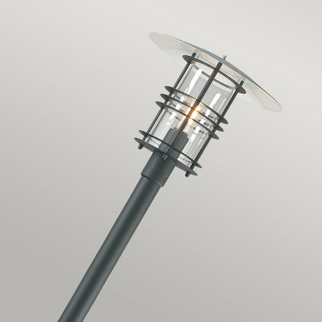 The Stockholm 1 Light Grande Lamp Post - Black features a sleek, modern design with a black pole and Scandinavian-inspired cylindrical glass enclosure that houses a lit bulb. Its flat, disc-shaped top elegantly stands out against a simple, light gray background, making it the epitome of chic outdoor lighting.