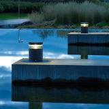 The Stavanger LED Small Bollards in graphite cast a sleek, modern glow along a concrete pathway above a reflective water surface, framed by tall grasses. The tranquil scene is beautifully captured at twilight, with the deep blue hues of the sky mirrored on the water.