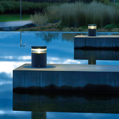 The Stavanger LED Small Bollards in graphite cast a sleek, modern glow along a concrete pathway above a reflective water surface, framed by tall grasses. The tranquil scene is beautifully captured at twilight, with the deep blue hues of the sky mirrored on the water.