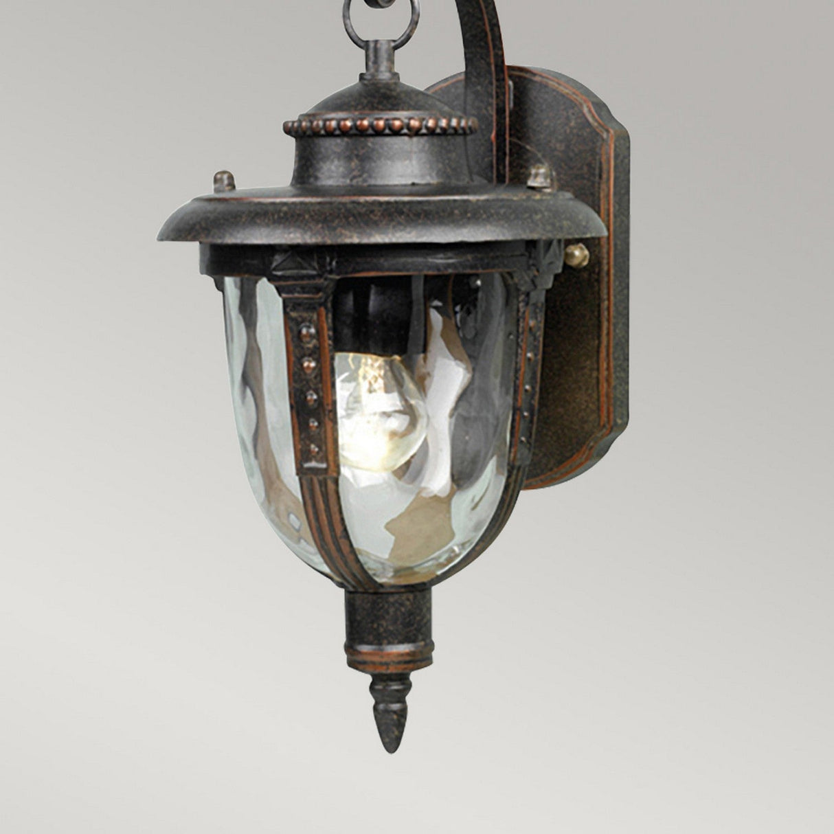 The St Louis 1 Light Outdoor Small Wall Light in weathered bronze finish, featuring glass panels, is mounted on a gray wall. With its ornamental design and visible bulb, it presents a classic and elegant look.