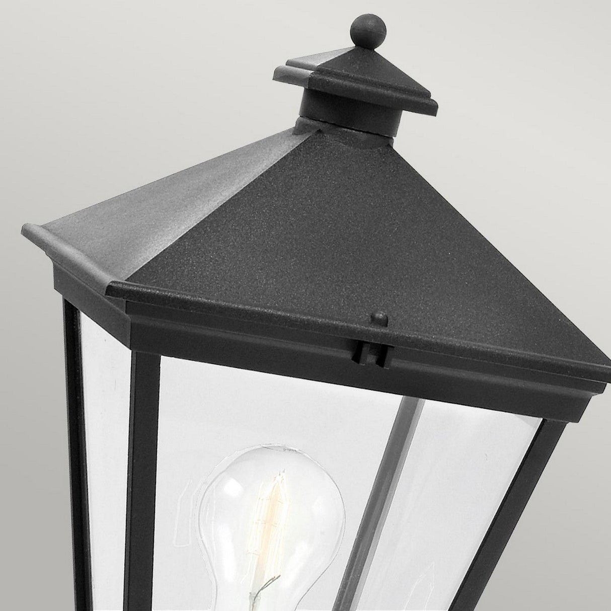A close-up of the Turin 1 Light Pedestal in black features a pyramid-shaped top and clear glass panels with a visible incandescent bulb inside. Made from die-cast aluminum, this elegant outdoor lighting piece comes with an IP54 rating, set against a neutral gray background.
