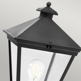 A close-up of the Turin 1 Light Pedestal in black features a pyramid-shaped top and clear glass panels with a visible incandescent bulb inside. Made from die-cast aluminum, this elegant outdoor lighting piece comes with an IP54 rating, set against a neutral gray background.