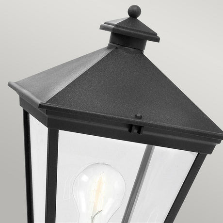 A close-up of the Turin 1 Light Pedestal in black features a pyramid-shaped top and clear glass panels with a visible incandescent bulb inside. Made from die-cast aluminum, this elegant outdoor lighting piece comes with an IP54 rating, set against a neutral gray background.