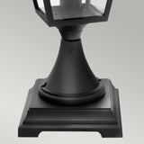 Close-up of the base of the Turin 1 Light Pedestal - Black, an outdoor lantern made from die-cast aluminum with a square, stepped design. It is mounted on a tapered stand, exuding a minimalist and modern aesthetic. The lantern is suitable for various weather conditions due to its IP54 rating, set against a plain gray background.