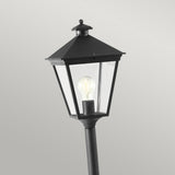 The Turin 1 Light Pillar - Black is a die-cast aluminium outdoor lantern-style lamp post featuring a pointed top and clear glass enclosure that reveals a visible bulb. Its classic design effortlessly complements outdoor landscapes against a simple gray backdrop.