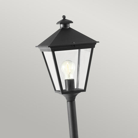The Turin 1 Light Pillar - Black is a die-cast aluminium outdoor lantern-style lamp post featuring a pointed top and clear glass enclosure that reveals a visible bulb. Its classic design effortlessly complements outdoor landscapes against a simple gray backdrop.