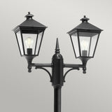 The Turin 2 Light Twin Lamp Post - Black features a black metal structure crafted from die-cast aluminum with clear glass panels that showcase visible light bulbs. It stands against a plain gray background, offering an IP54 rating and a classic angular design reminiscent of a vintage style.