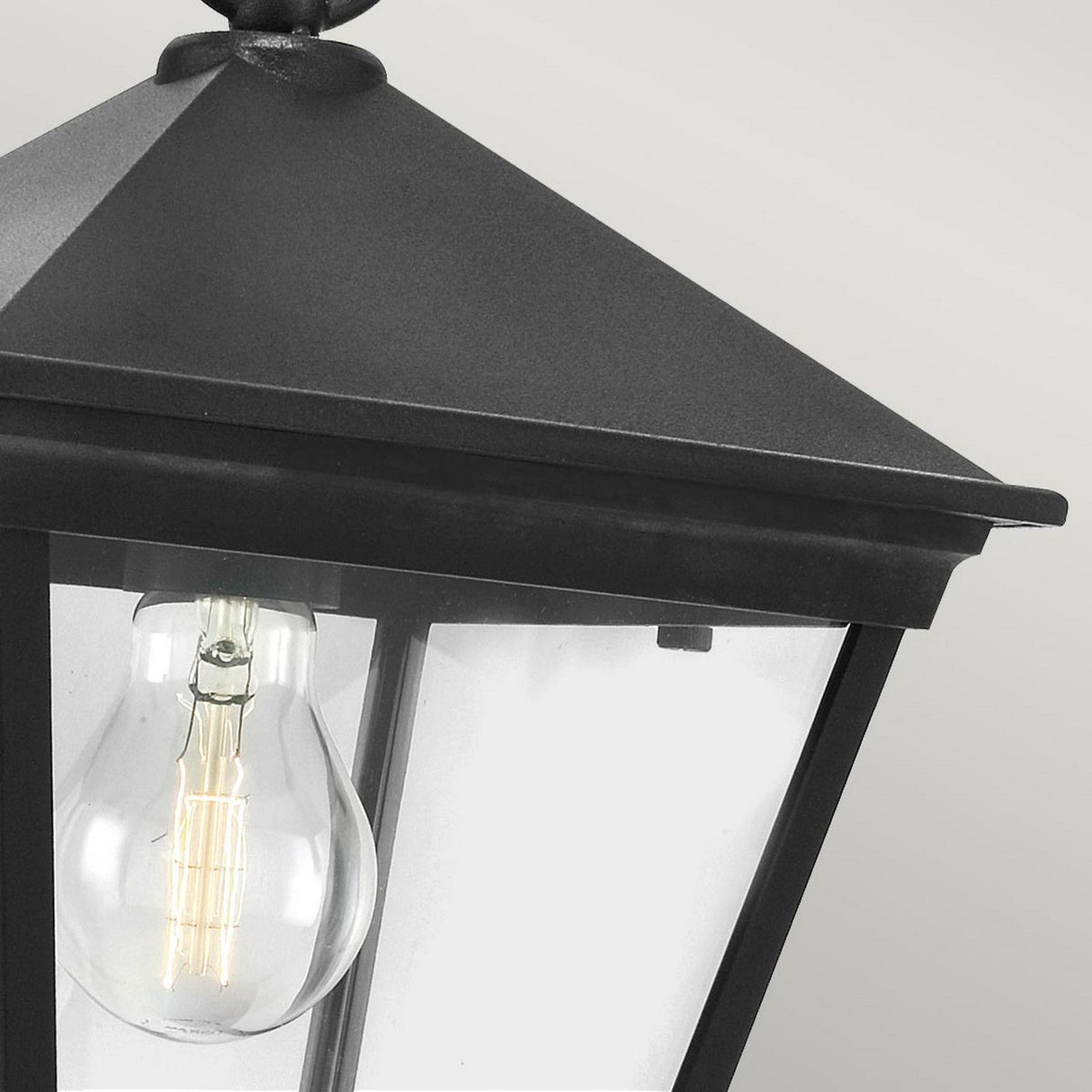 A close-up of the Turin Outdoor 1 Light Chain Pendant in black showcases its transparent glass cover and a glowing light bulb inside. This weather-resistant lighting fixture boasts a classic pyramid-shaped top, hanging elegantly from a black chain against a light gray background.