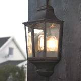 The Turin Outdoor 1 Light Half Lantern – Black is a vintage-style outdoor lantern made from die-cast aluminum, boasting a black metal frame and clear glass panels. Rated IP54, it emits a soft, warm light when installed on a dark wooden surface, with the blurred background of house and sky enhancing its charm.