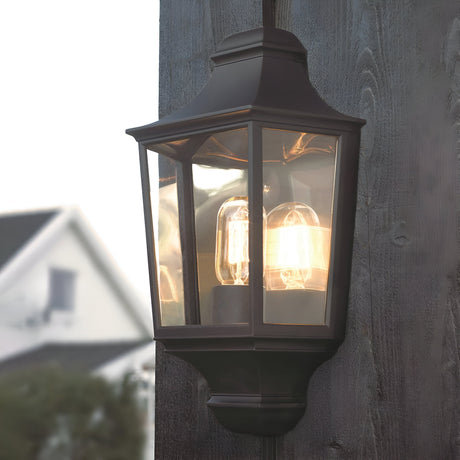 The Turin Outdoor 1 Light Half Lantern – Black is a vintage-style outdoor lantern made from die-cast aluminum, boasting a black metal frame and clear glass panels. Rated IP54, it emits a soft, warm light when installed on a dark wooden surface, with the blurred background of house and sky enhancing its charm.