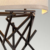 A close-up reveals a modern table lamp with a geometric metal frame finished in Olde Bronze, showcasing intersecting rods. The slightly curved light beige lampshade emits a warm glow ideal for decorative lighting. Set against a neutral gray background, it perfectly complements the style of the Taiko 2 Light Wall Light.